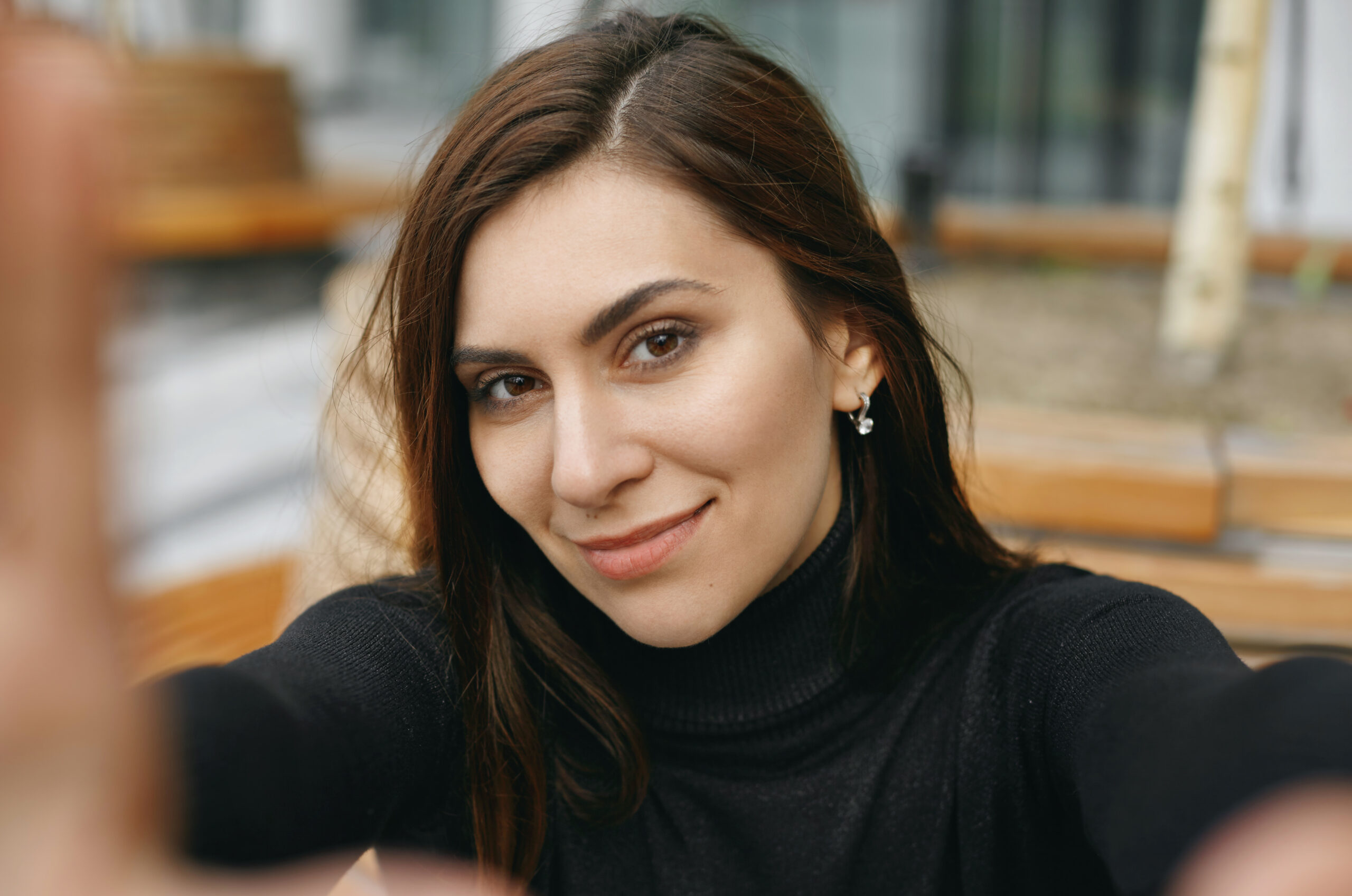 outdoor portrait of pretty young brunette female taking selfie flirty smiling at camera. charming girl looking at camera with happy joyful expression and gesturing hands frame. selective focus on face